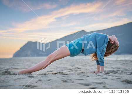 A woman practices Purvottanasana, or upward plank pose, on the beach during sunset. The golden sky reflects on her focused core work as she balances on the sand in serene surroundings. 133413506