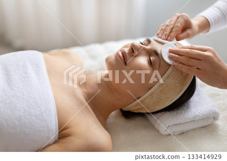 Therapist cleansing forehead of young beautiful woman with cotton pads. Therapist cleansing forehead of young beautiful woman with cotton pads. 133414929