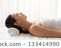 Young woman wrapped in towel lying on spa bed with eyes closed 133414990