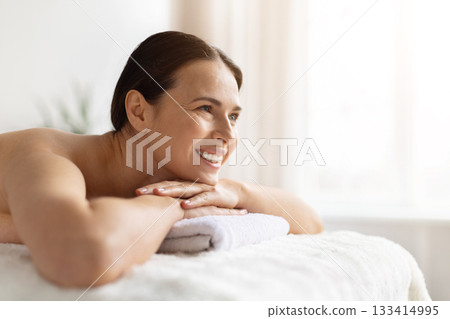 Smiling young woman lying on spa bed, resting her chin on towel Smiling young woman lying on spa bed, resting her chin on towel 133414995
