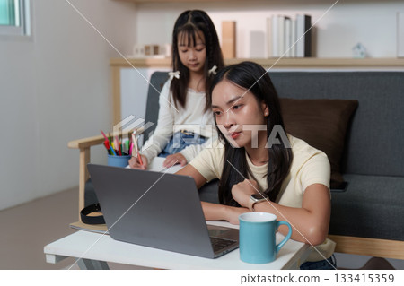 Remote Learning. Young woman studying on a laptop while younger girl draws. 133415359