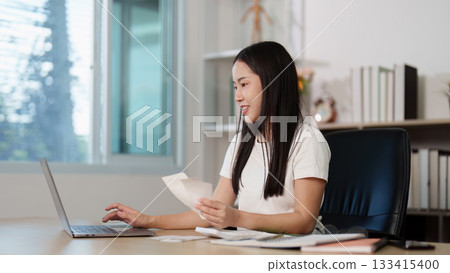 Remote Work. Young woman managing finances at home office with laptop and documents. 133415400