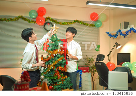 Happy two young businessman decorating festive Christmas tree together in modern office. Woman smiling while hanging red ornament 133415936