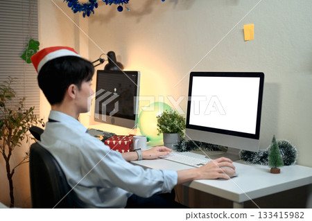 Tired man in Santa hat working and using a computer white screen at the working desk overtime Tired man in Santa hat working and using a computer white screen at the working desk overtime 133415982