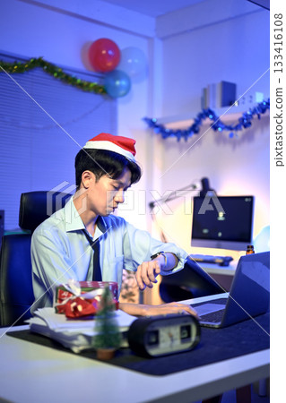 Businessman in Santa hat working late at Christmas day in office with Christmas decor 133416108