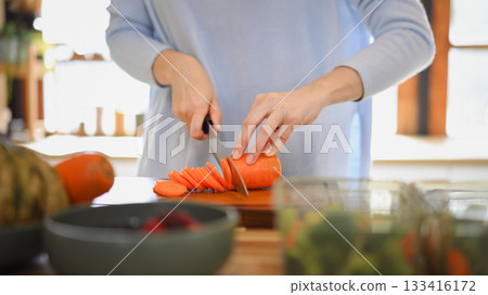 Closeup of young female hands chopping fresh orange carrot on board while in modern kitchen Closeup of young female hands chopping fresh orange carrot on board while in modern kitchen 133416172