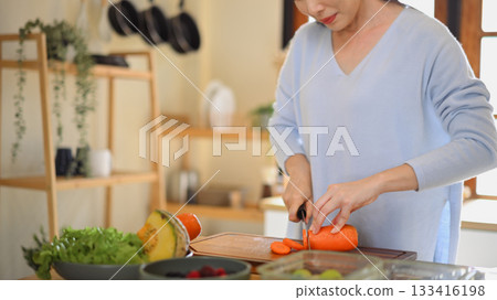 Closeup of young female hands chopping fresh orange carrot on board while in modern kitchen Closeup of young female hands chopping fresh orange carrot on board while in modern kitchen 133416198