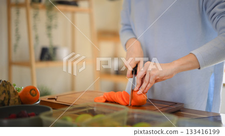 Closeup of young female hands chopping fresh orange carrot on board while in modern kitchen Closeup of young female hands chopping fresh orange carrot on board while in modern kitchen 133416199