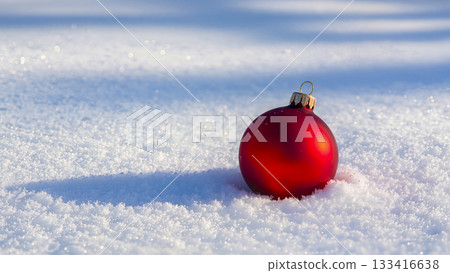 A red Christmas tree ball in the snow. A festive decoration on a fluffy blanket of snow on a bright sunny day with long shadows. 133416638