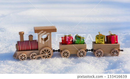 A wooden toy train with presents in the snow. A Christmas locomotive with carriages loaded with brightly colored boxes. 133416639