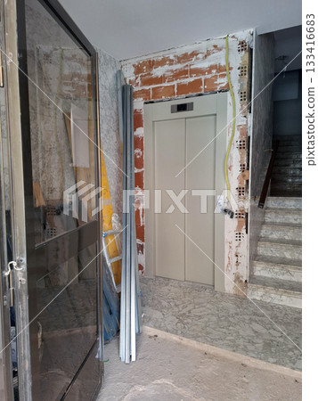 Workers are replacing the old elevator at the entrance of the building. Construction materials and tools are scattered around the area. The nearby stairs remain accessible during the work. 133416683