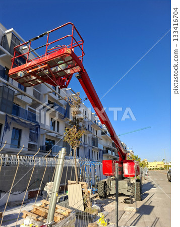 A bright red painter's lift stands tall against a clear blue sky at a construction site. Workers use the lift to reach higher areas of the building nearing completion. 133416704