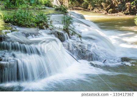 A small waterfall cascades over layered rocks surrounded by trees, with the water flowing in shallow steps into a wider pool. 133417367
