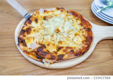 A person uses a knife to cut a slice of thin crust pizza on a round wooden board, showing melted cheese and pineapple pieces on top. A person uses a knife to cut a slice of thin crust pizza on a round wooden board, showing melted cheese and pineapple pieces on top. 133417369