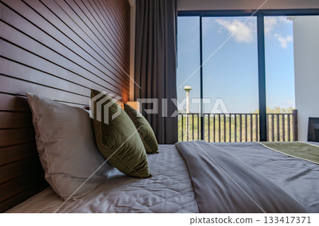 A bed with white sheets and green pillows sits against a wooden panel wall, with two wall lamps and small side tables creating a simple layout. A bed with white sheets and green pillows sits against a wooden panel wall, with two wall lamps and small side tables creating a simple layout. 133417371