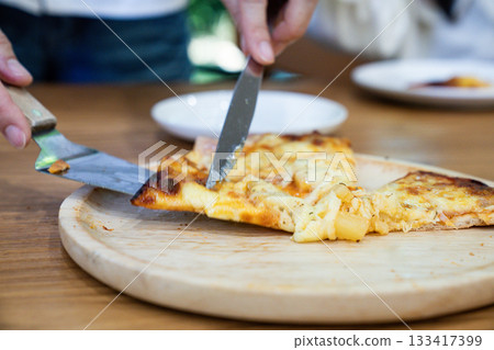 A person uses a knife to cut a slice of thin crust pizza on a round wooden board, showing melted cheese and pineapple pieces on top. 133417399