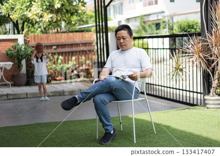 A man reads peacefully while a young girl holds a basketball in the background. A soft moment showing connection across generations at home. A man reads peacefully while a young girl holds a basketball in the background. A soft moment showing connection across generations at home. 133417407