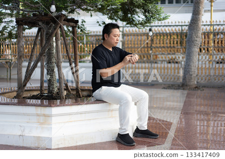 A man sits quietly in the temple courtyard, checking the time on his smartwatch while waiting calmly in the peaceful surroundings. 133417409