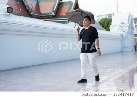 A man walks calmly with an umbrella near a temple wall, reflecting a peaceful moment after rainfall with a relaxed and mindful expression. 133417417