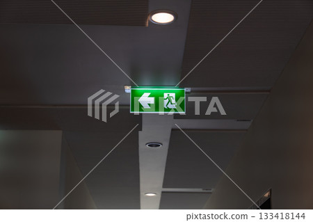 A green fire escape sign hangs from the ceiling, showing an exit sign green light box with a running man and an arrow pointing right for safety and evacuation. A green fire escape sign hangs from the ceiling, showing an exit sign green light box with a running man and an arrow pointing right for safety and evacuation. 133418144