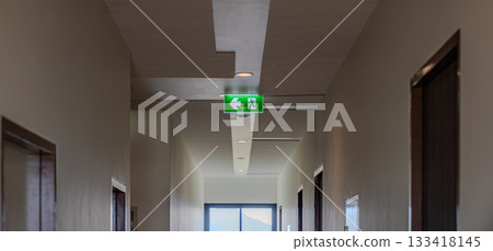 A green fire escape sign hangs from the ceiling, showing an exit sign green light box with a running man and an arrow pointing right for safety and evacuation. A green fire escape sign hangs from the ceiling, showing an exit sign green light box with a running man and an arrow pointing right for safety and evacuation. 133418145