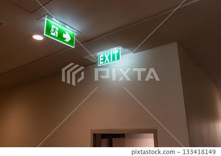 A green fire escape sign hangs from the ceiling, showing an exit sign green light box with a running man and an arrow pointing right for safety and evacuation. A green fire escape sign hangs from the ceiling, showing an exit sign green light box with a running man and an arrow pointing right for safety and evacuation. 133418149