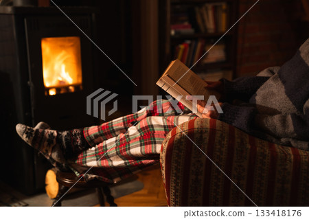 A woman reads a book while sitting in a chair next to a stove in the evening. 133418176