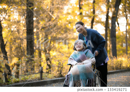 一位女士在落葉紛飛的仲秋時節，在戶外照顧一位坐在輪椅上的老人。這幅畫面展現了外出居家照護或散步的情景。 133419271