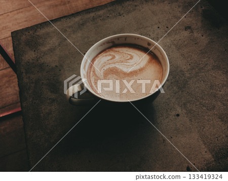 Cup of hot coffee with latte art on a rustic stone table. Warm and cozy cafe atmosphere, close-up of cappuccino with frothy milk on concrete table. 133419324
