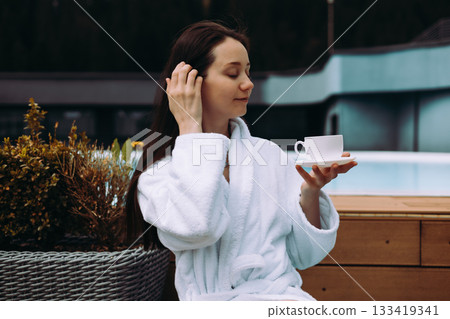 A relaxed woman enjoys cup of tea near a pool, wearing a white robe in a peaceful setting. A serene girl enjoys a moment with a cup of morning coffee. A relaxed woman enjoys cup of tea near a pool, wearing a white robe in a peaceful setting. A serene girl enjoys a moment with a cup of morning coffee. 133419341