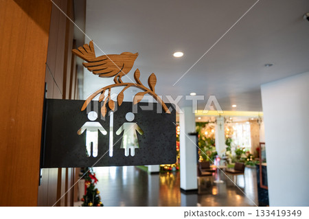 A black steel restroom sign with male and female symbols hangs on a wall. The cutout icons are placed under a carved bird and branch decoration. A black steel restroom sign with male and female symbols hangs on a wall. The cutout icons are placed under a carved bird and branch decoration. 133419349