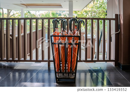 Closed orange umbrellas arranged neatly in a black metal stand, with outside light highlighting the folded fabric and curved handles. Closed orange umbrellas arranged neatly in a black metal stand, with outside light highlighting the folded fabric and curved handles. 133419352