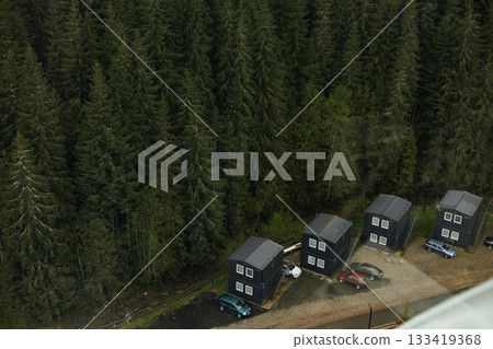 Aerial shot of small black wooden cabins nestled near a dense coniferous forest, surrounded by lush green pine trees and parked cars on the road side. Aerial shot of small black wooden cabins nestled near a dense coniferous forest, surrounded by lush green pine trees and parked cars on the road side. 133419368
