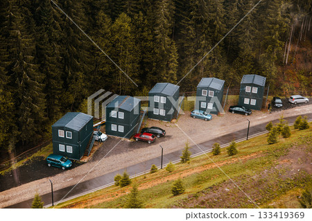 Aerial shot of small black wooden cabins nestled near a dense coniferous forest, surrounded by lush green pine trees and parked cars on the road side. 133419369