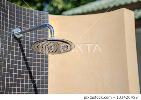 A metal shower head is mounted on a tiled wall with beige curved panels around it, and soft sunlight highlights the tile texture and the metal surface. 133420459