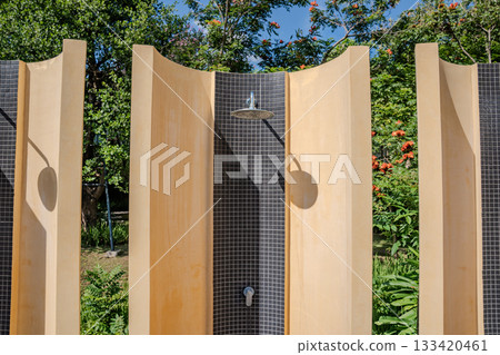 A metal shower head is mounted on a tiled wall with beige curved panels around it, and soft sunlight highlights the tile texture and the metal surface. A metal shower head is mounted on a tiled wall with beige curved panels around it, and soft sunlight highlights the tile texture and the metal surface. 133420461