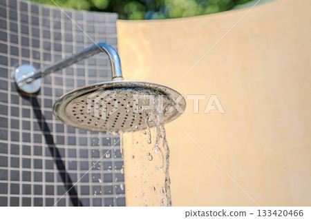Water flows from a round metal shower head against a tiled wall, with sunlight highlighting the water texture and the outdoor setting. Water flows from a round metal shower head against a tiled wall, with sunlight highlighting the water texture and the outdoor setting. 133420466