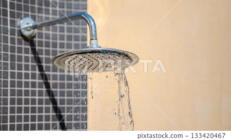 Water flows from a round metal shower head against a tiled wall, with sunlight highlighting the water texture and the outdoor setting. 133420467