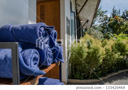 Rolled blue towels are stacked on a wooden shelf beside the pool walkway, with sunlight highlighting the towel texture and the surrounding plants. 133420469