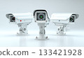 Three white modern security cameras with multiple small bright lights stand on a clean reflective surface representing advanced digital protection and continuous watchful monitoring. 133421928