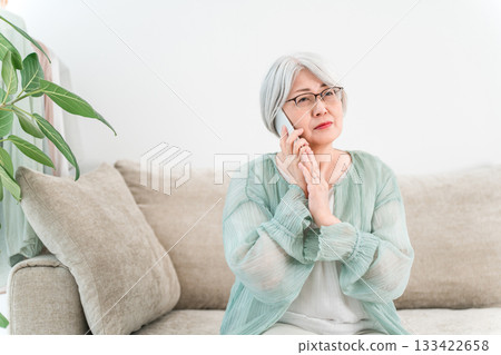 A gray-haired senior woman with a troubled expression while talking on her smartphone (scam call, suspicious call, nuisance call) 133422658
