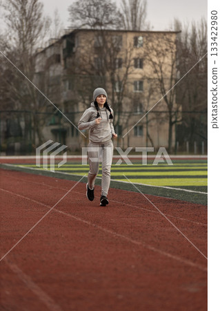 female track and field athlete jogs on a red running track at a sports facility. She wears comfortable athletic clothing and a beanie showing focus and determination in her training session. female track and field athlete jogs on a red running track at a sports facility. She wears comfortable athletic clothing and a beanie showing focus and determination in her training session. 133422980