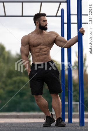 athletic man stands confidently on a sports ground showcasing his muscular physique while resting against a bar. The environment is ideal for outdoor workouts and fitness training. 133422988