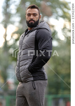 athletic man stands on a sports ground dressed in a warm jacket and workout pants. The scene captures the early morning light as he prepares for an active session. athletic man stands on a sports ground dressed in a warm jacket and workout pants. The scene captures the early morning light as he prepares for an active session. 133422994