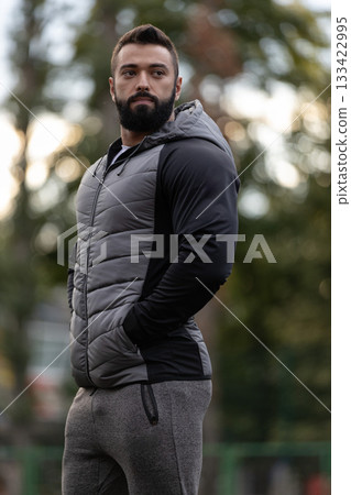 athletic man wearing a gray vest and sweatpants stands with his hands in pockets on a sports ground. The setting sun creates a warm atmosphere around him in the evening light. 133422995