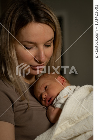A nurturing mother cradles her peaceful baby in her arms, creating a loving bond in the comfort of their cozy home during a calm evening. 133423003