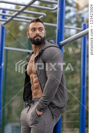 muscular man is confidently posing on a sports ground showcasing his toned physique while dressed in a gray athletic outfit. The setting is bright and inviting. 133423004