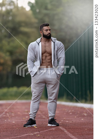 muscular man stands on a running track wearing athletic clothing. He appears focused and relaxed highlighting his fitness journey. The background features trees and a clear sky. muscular man stands on a running track wearing athletic clothing. He appears focused and relaxed highlighting his fitness journey. The background features trees and a clear sky. 133423020