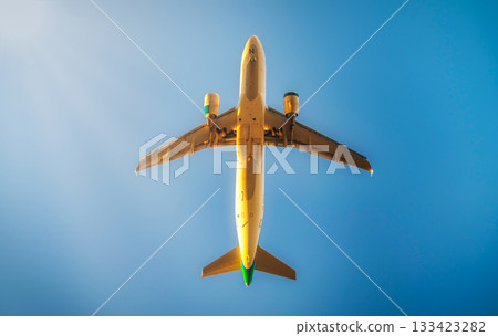 The image shows an airplane captured from below, with its underbelly and landing gear clearly visible against a clear blue sky. The sunlight is casting a glow on parts of the plane, creating a few The image shows an airplane captured from below, with its underbelly and landing gear clearly visible against a clear blue sky. The sunlight is casting a glow on parts of the plane, creating a few 133423282