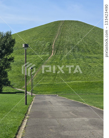 A paved path leads toward a large grassy hill with a walking trail, creating a sense of journey and exploration A paved path leads toward a large grassy hill with a walking trail, creating a sense of journey and exploration 133423490